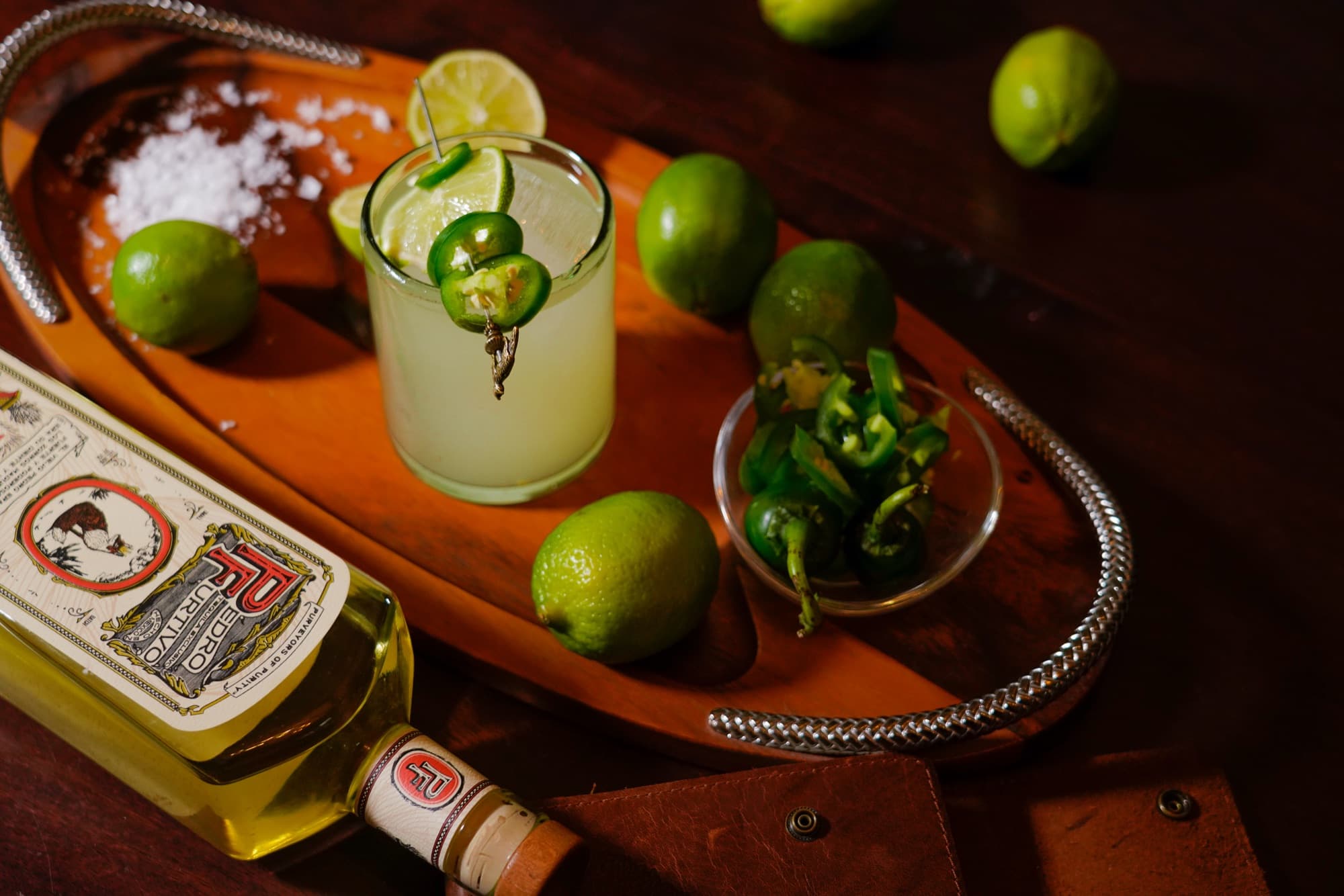 A Jalapeño Margarita on a wooden serving tray with limes, coarse salt, a bowl of sliced jalapeños and a bottle of Pedro Furtivo Reposado