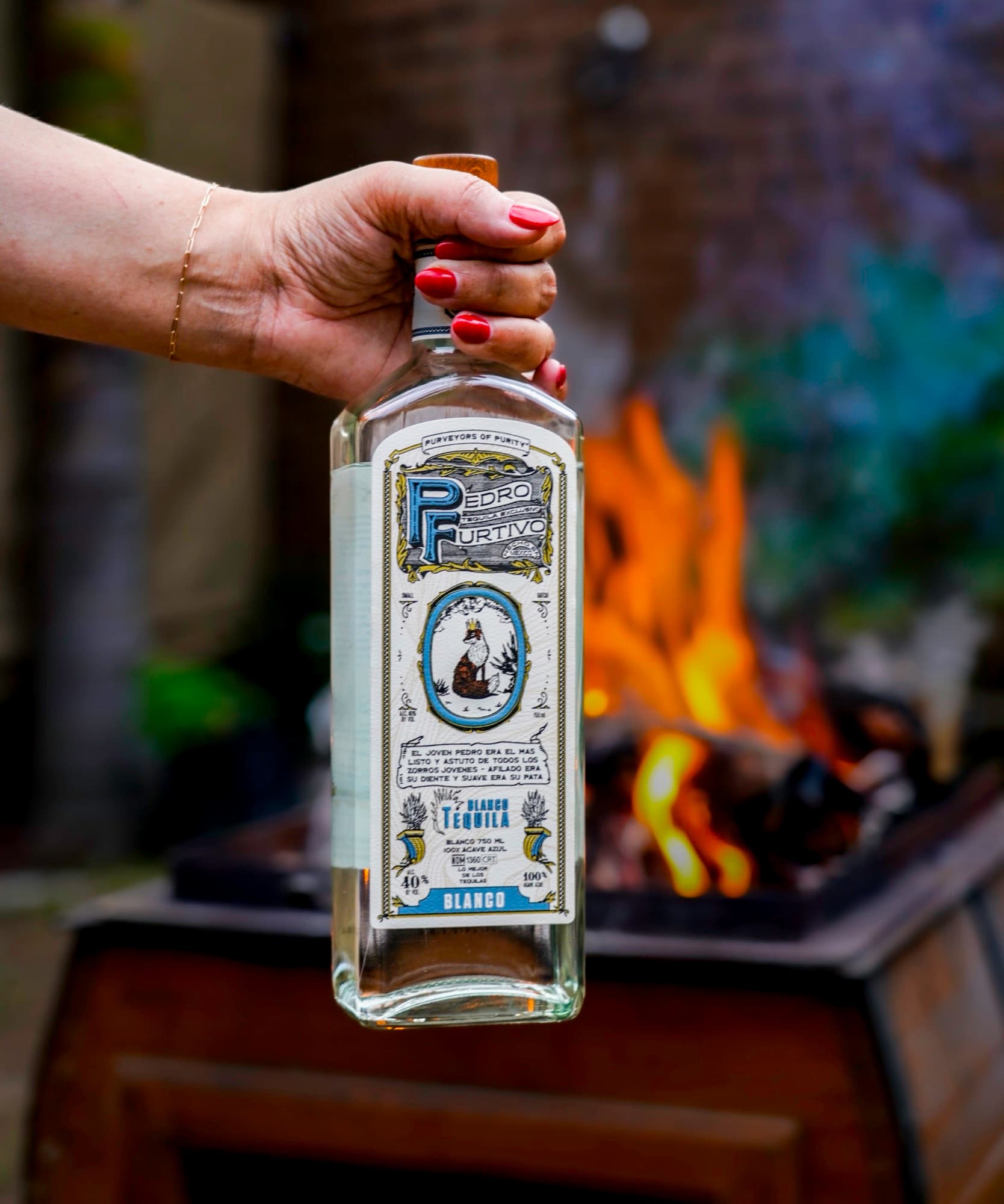 A hand lifting a bottle of Pedro Furtivo Blanco in front of an open fire