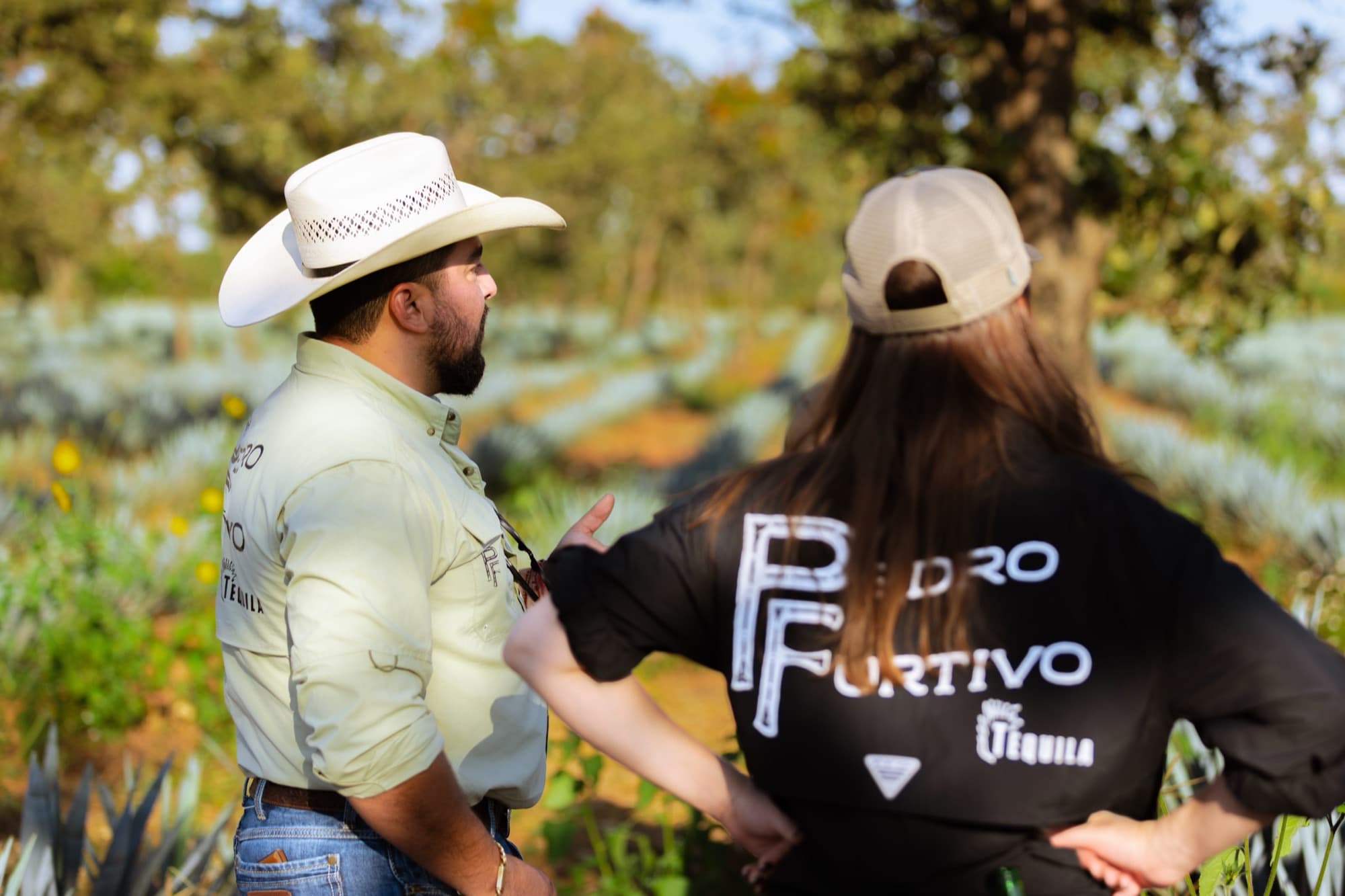 A woman in a black Pedro Furtivo tee and cap, seen from behind, talking with a man in a cowboy hat among the agave
