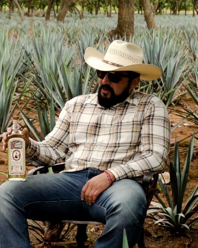 Omar Martinez in a plaid shirt and straw hat, seated in the agave rows with a bottle of Pedro Furtivo beside him