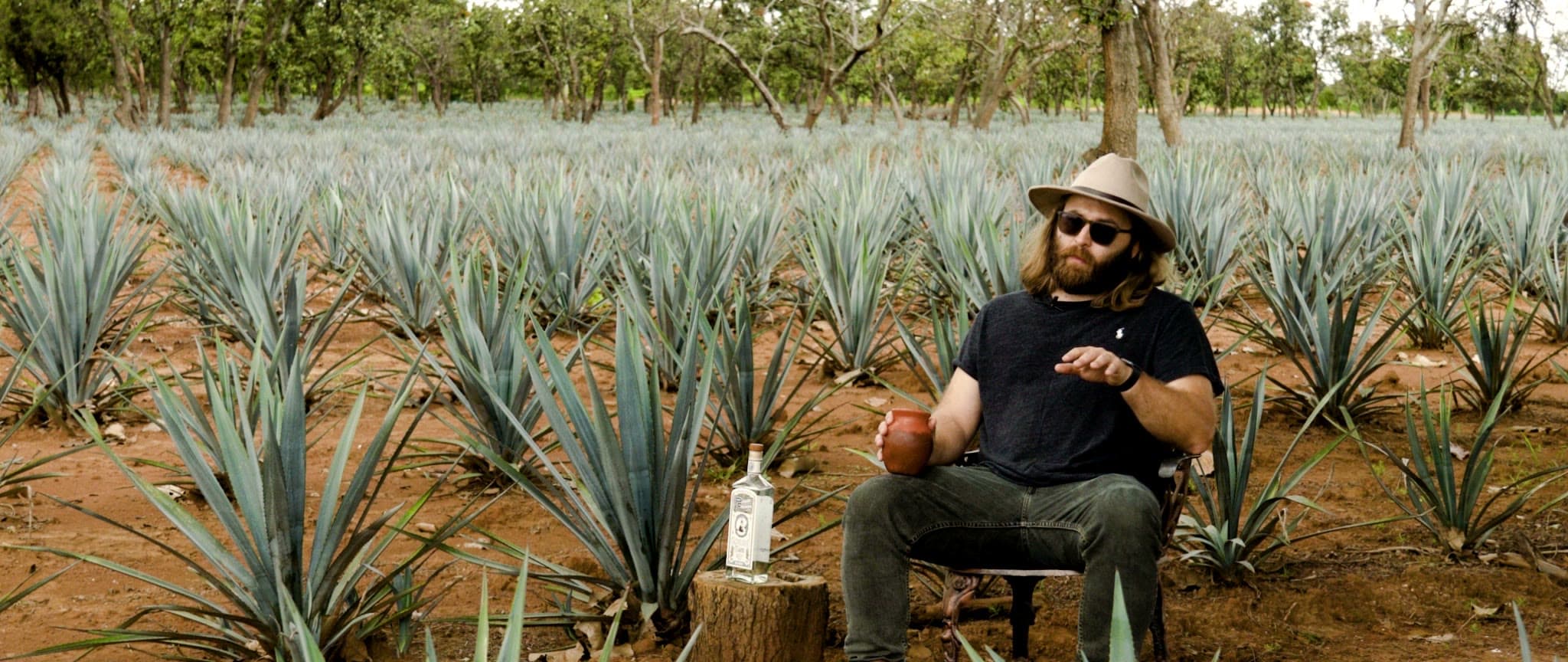 Peter Kiley seated among the agave rows in a black tee and hat, holding a clay cup, a bottle of Pedro Furtivo on a stump beside him, from the Origin Story film