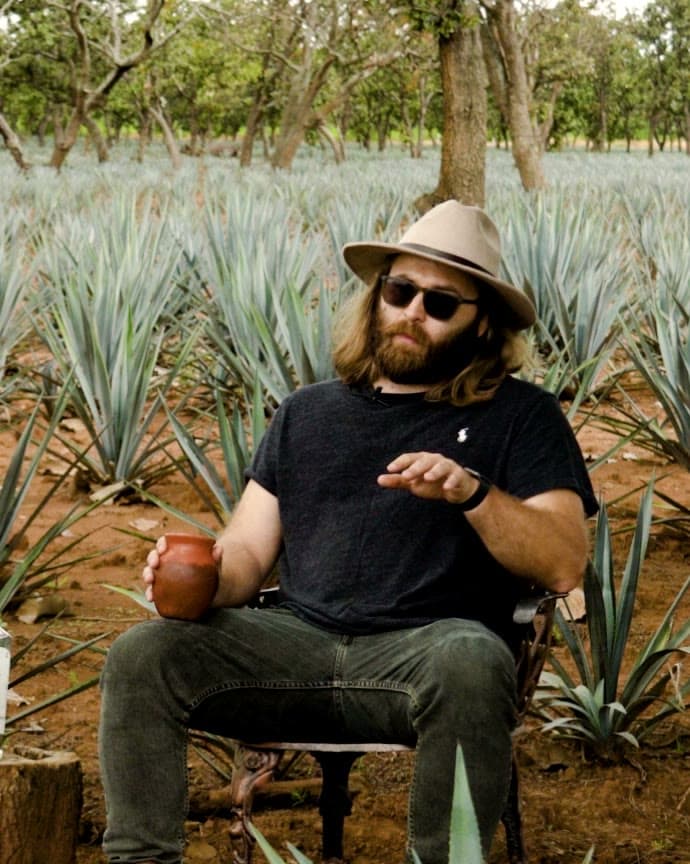 Peter Kiley in a black tee and hat, seated in the agave rows with a clay cup
