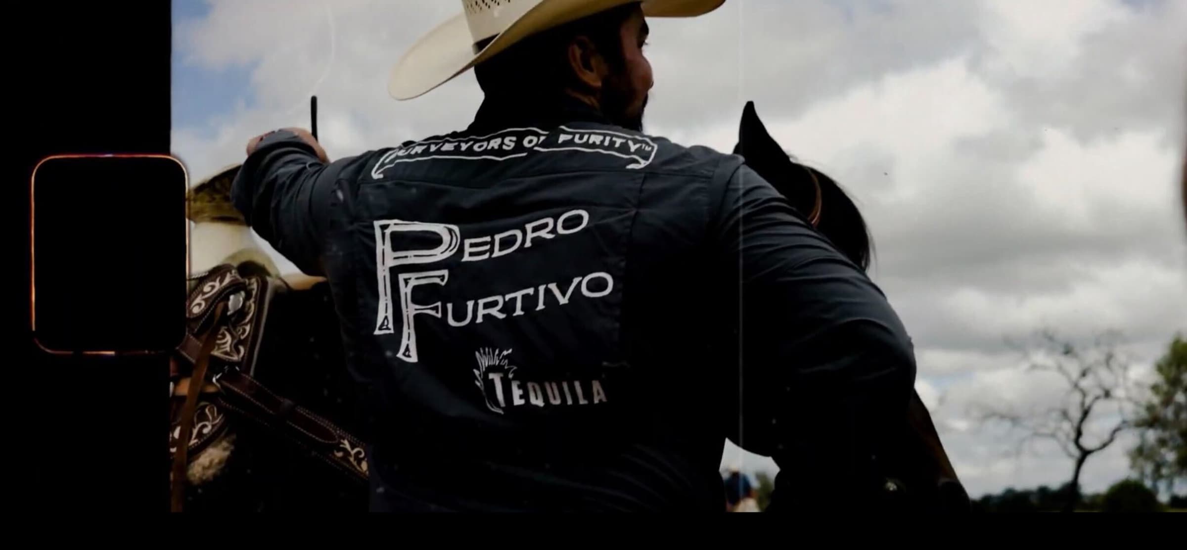 The back of a black Pedro Furtivo work shirt, Purveyors of Purity across the shoulders, worn by a man in a cowboy hat under a cloudy sky