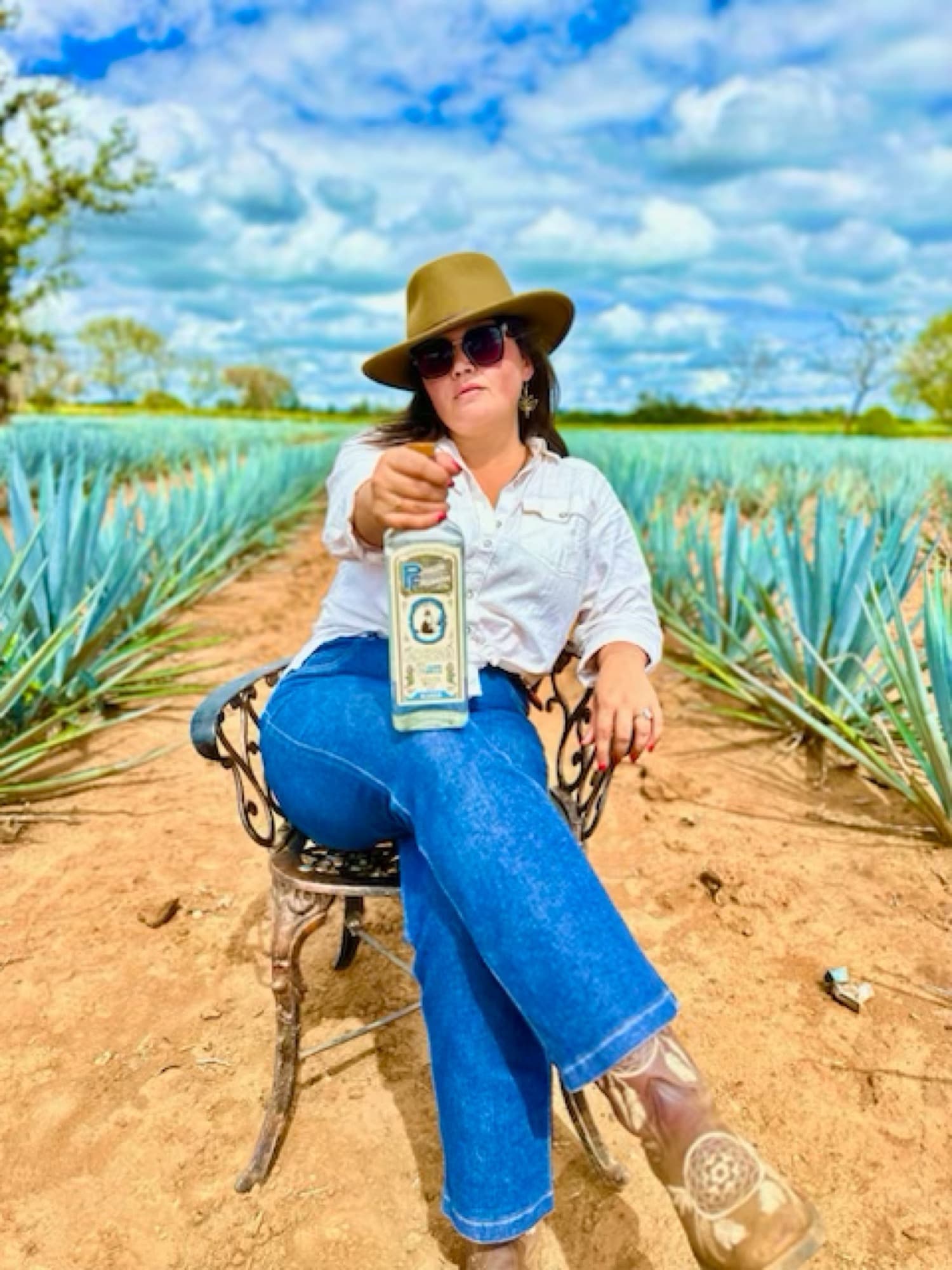 Rachel Kiley in a wide-brimmed hat, seated among rows of blue agave and holding out a bottle of Pedro Furtivo