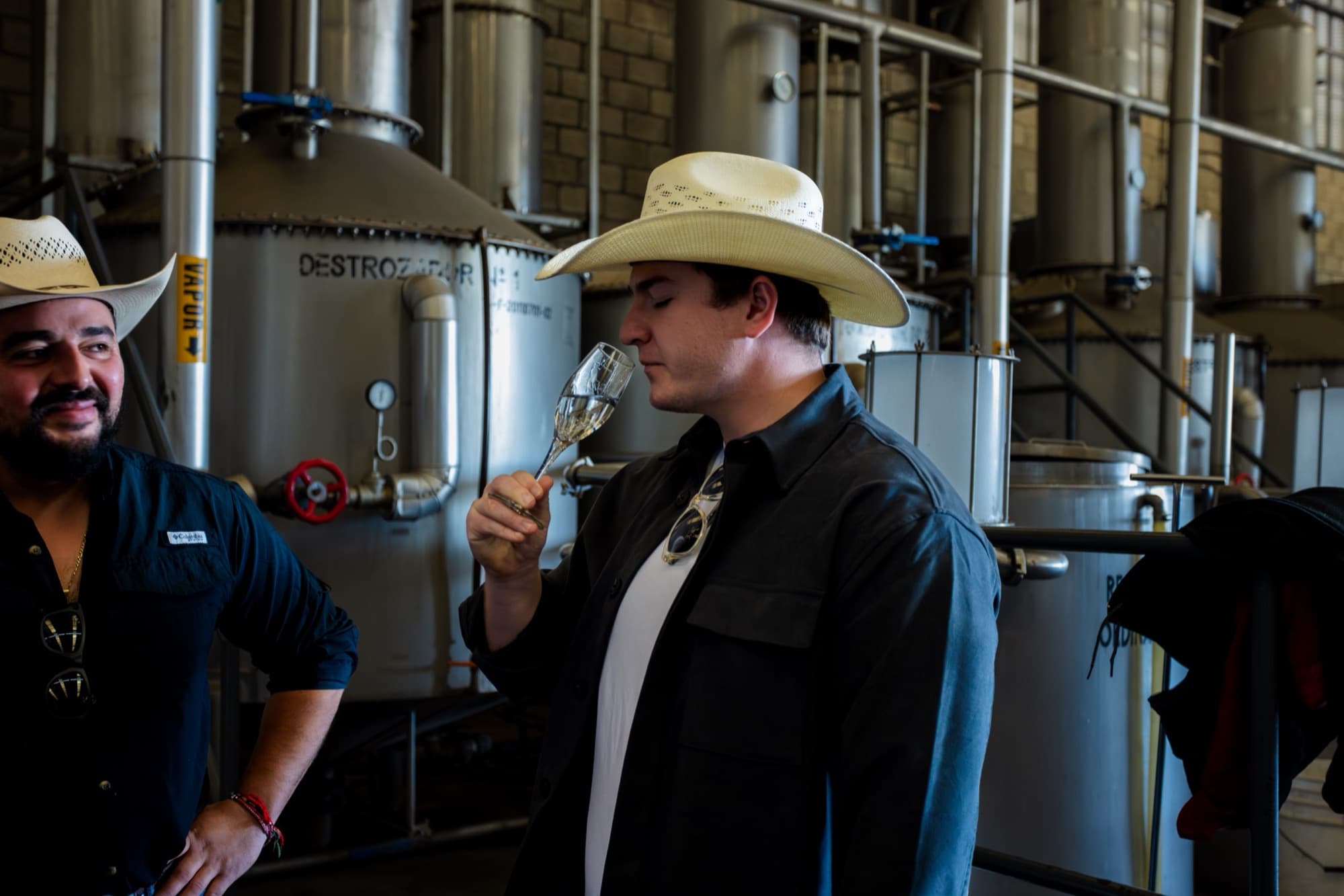 Two men tasting tequila beside the stainless-steel stills
