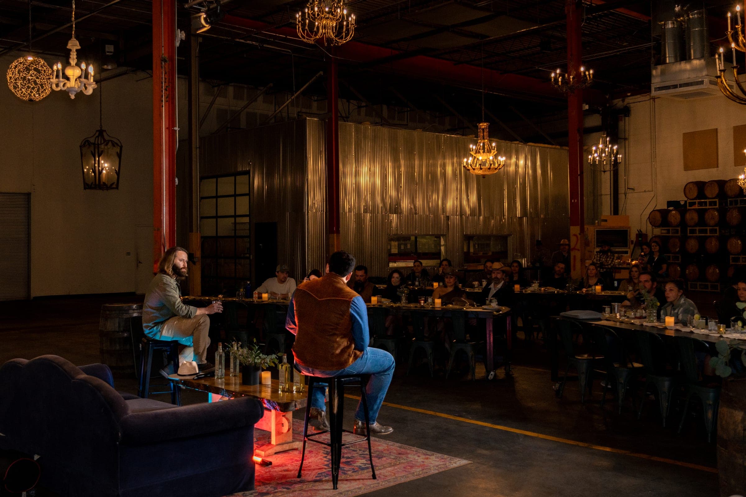 Peter Kiley and Omar Martinez on stage stools, talking to a candlelit room of guests under chandeliers at the Origin Story premiere party