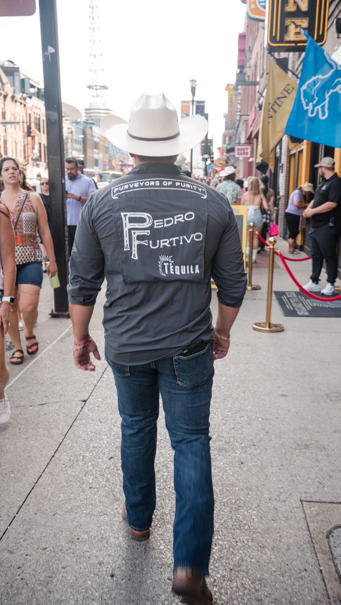 A man in a white cowboy hat walking down a busy sidewalk, Purveyors of Purity and Pedro Furtivo Tequila printed across the back of his charcoal shirt