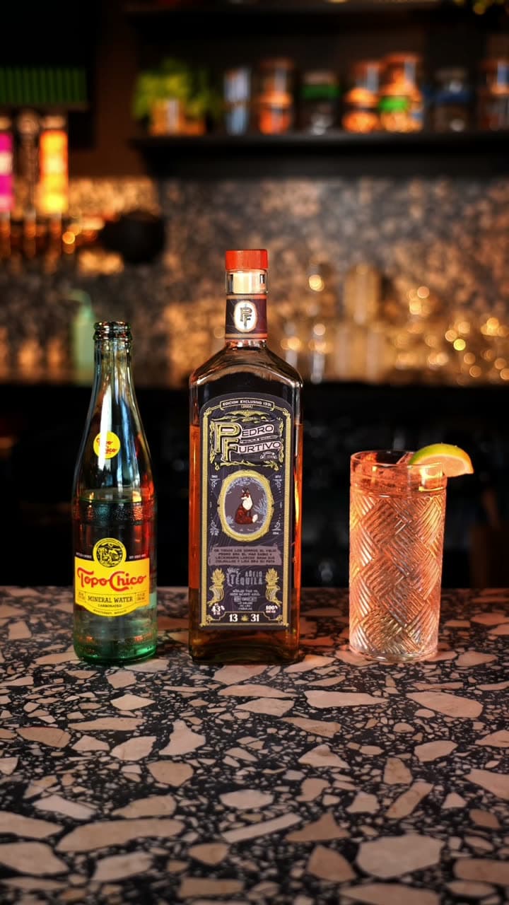 A bottle of Pedro Furtivo 1331 Añejo beside a Topo Chico and a tall highball with a lemon wheel on a terrazzo bar