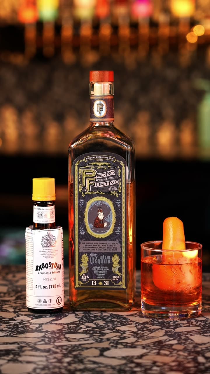 A bottle of Pedro Furtivo 1331 Añejo beside Angostura bitters and an old fashioned with an orange peel on a terrazzo bar