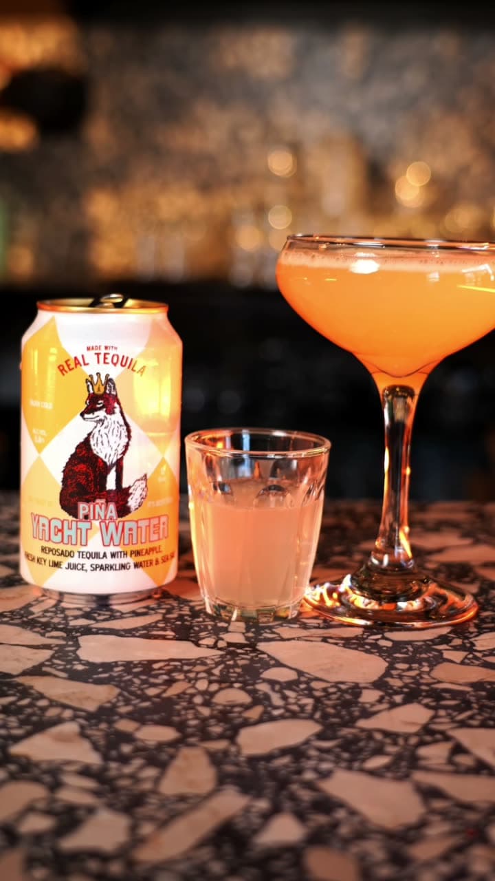 A pale orange cocktail in a coupe beside a can of Piña Tight Water and a small glass on a terrazzo bar
