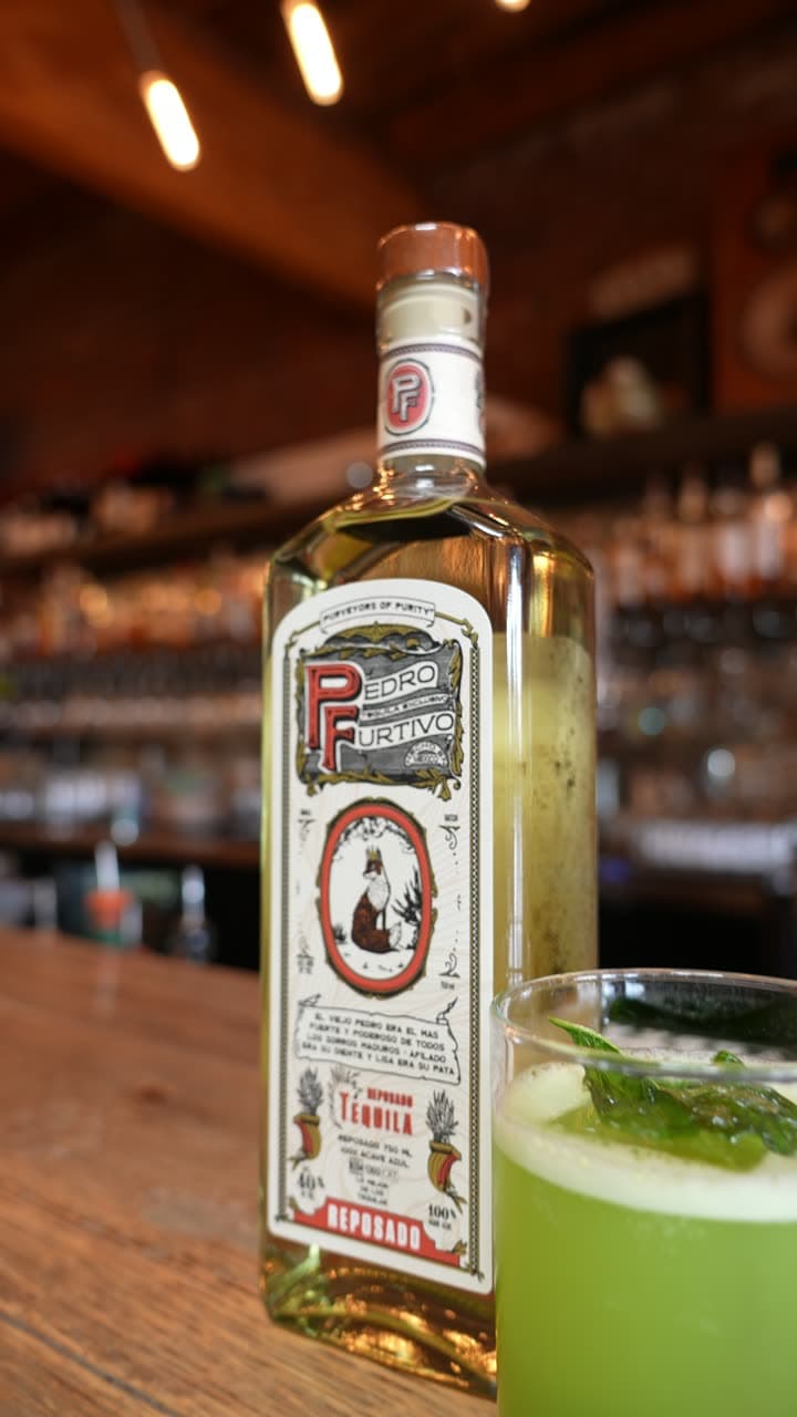 Pedro Furtivo Reposado beside a green cucumber cocktail on the bar
